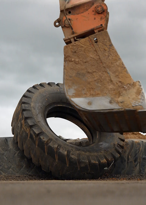 Close-up of a muddy excavator bucket lifting a large, dirty tire at a construction site under cloudy sky.
