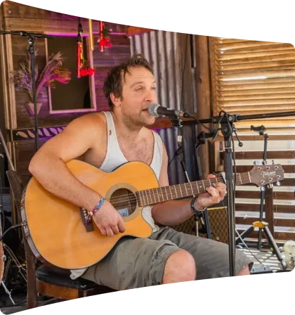 Man wearing a sleeveless shirt and shorts playing an acoustic guitar and singing into a microphone indoors.