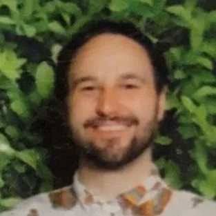 Smiling man with dark hair and beard wearing a patterned shirt, standing in front of green leafy background.