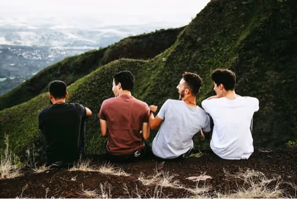 Four young men sitting on a grassy hillside with their backs to the camera, overlooking a vast landscape with hills and valleys in the distance, sharing a moment of laughter.