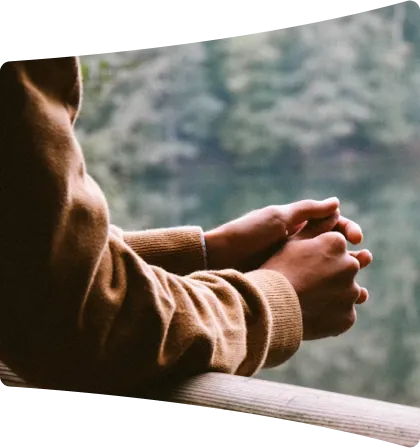 Person in a brown sweater leaning on a wooden railing with hands clasped, overlooking a calm body of water and a blurred green forest.