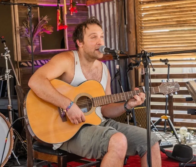 Man wearing a sleeveless shirt and shorts playing an acoustic guitar and singing into a microphone while seated on a wooden chair on a stage with musical instruments and equipment around him.