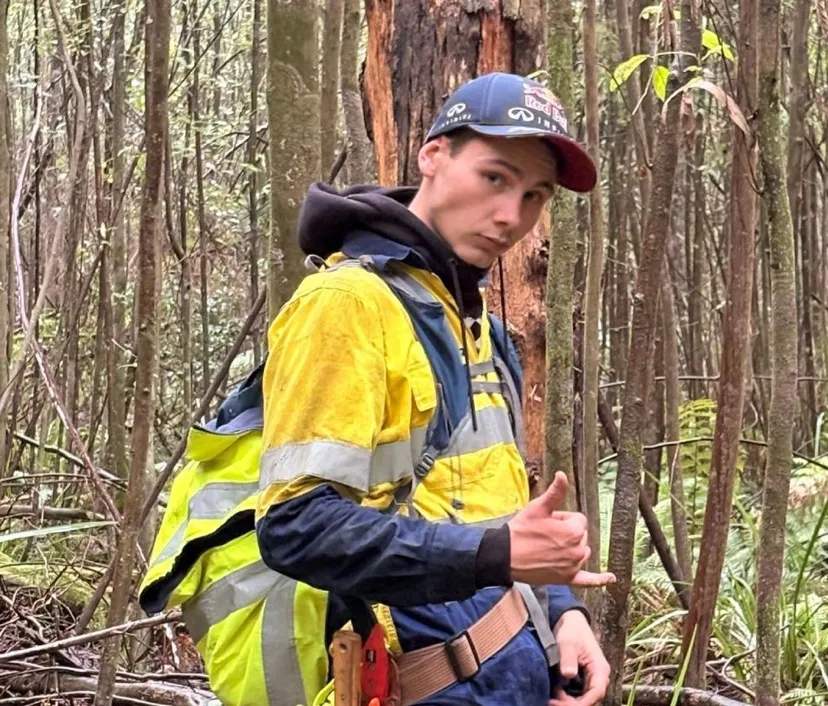 Young man wearing a yellow high-visibility jacket and a blue and red cap stands in a forest, making a shaka hand gesture.