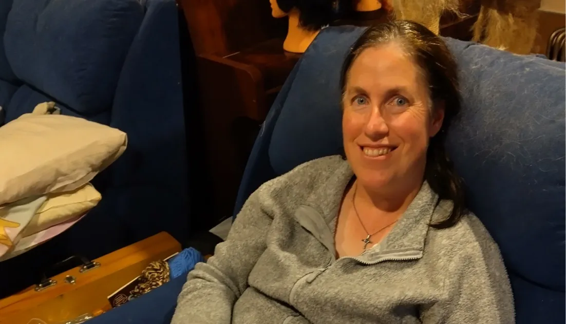 Smiling woman with light skin and dark hair wearing a gray fleece jacket sitting on a blue cushioned chair indoors.