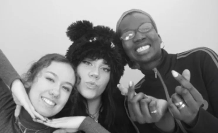 Three smiling friends posing together, one with hands under chin, one wearing a furry hat with ears, and one making a heart gesture with fingers.