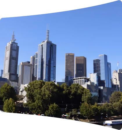 City skyline featuring modern high-rise buildings behind a large area of green trees under a clear blue sky.