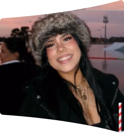 Smiling young woman with long dark hair wearing a furry hat and black jacket outdoors at dusk with another person in the background.