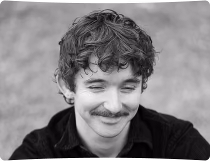 Black and white portrait of a young man with curly hair and a mustache looking down with a slight smile.