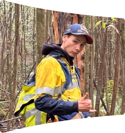 Young man wearing a yellow and black safety jacket and a cap making a hang loose hand gesture in a forest setting with tall trees.