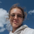 Person wearing sunglasses and a light-colored hoodie smiling against a blue sky with clouds.