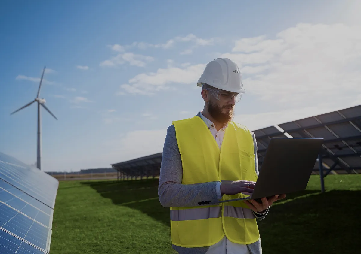Engineer in a white hard hat and yellow safety vest using a laptop in a solar panel field with a wind turbine in the background.