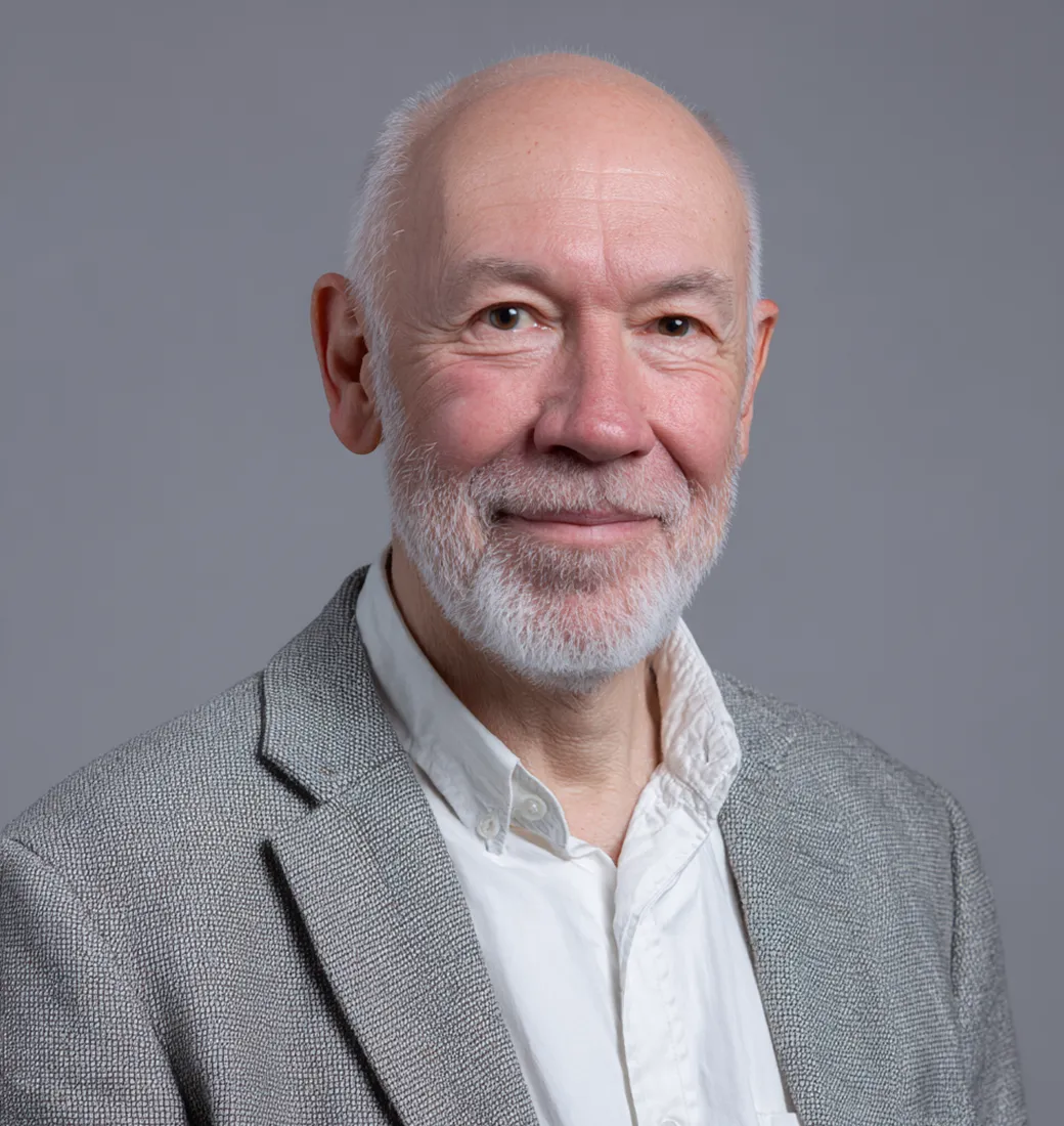 Portrait of an older bald man with a white beard wearing a gray blazer and white button-up shirt against a plain gray background.