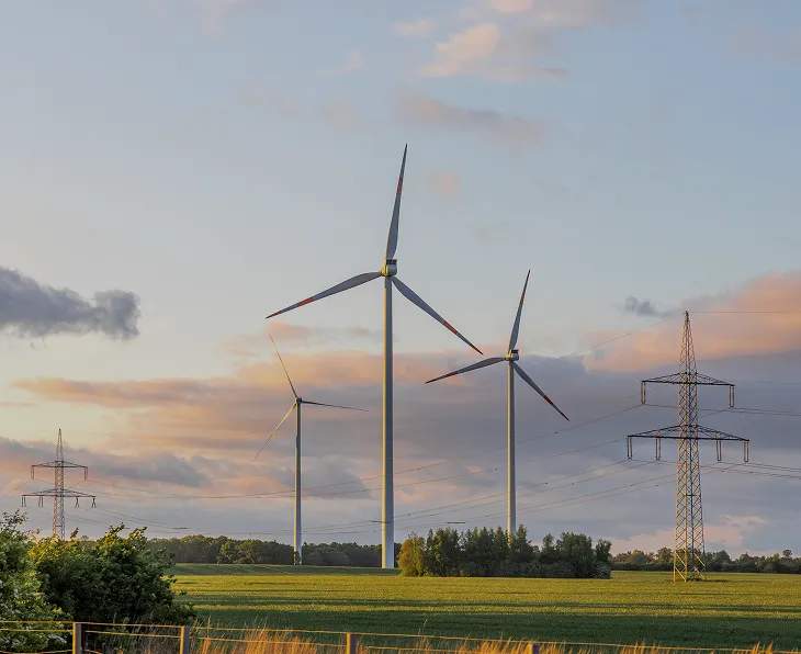 Three large wind turbines standing in a green field with power transmission towers and lines nearby under a partly cloudy sky at sunset.