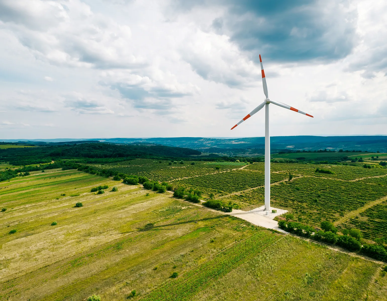 A large wind turbine with red-tipped blades stands in a green, open agricultural field under a partly cloudy sky, with hills in the distance.