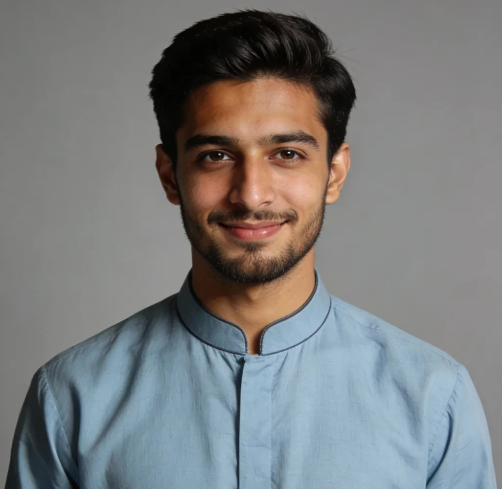 Young man with dark hair and beard smiling slightly, wearing a light blue traditional collarless shirt, against a plain gray background.