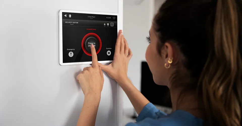Woman using a wall-mounted smart security system keypad, pressing the center button to disarm an alarm indicated by a glowing red circle on the screen.