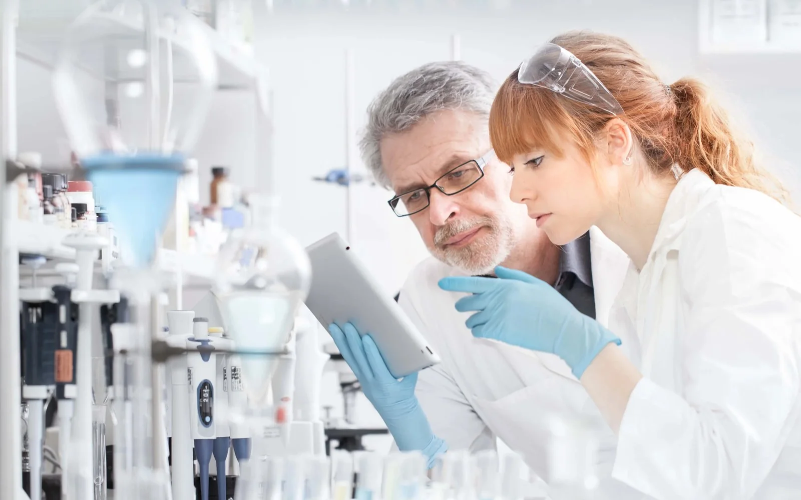 Two laboratory researchers reviewing scientific information on a tablet beside laboratory equipment.