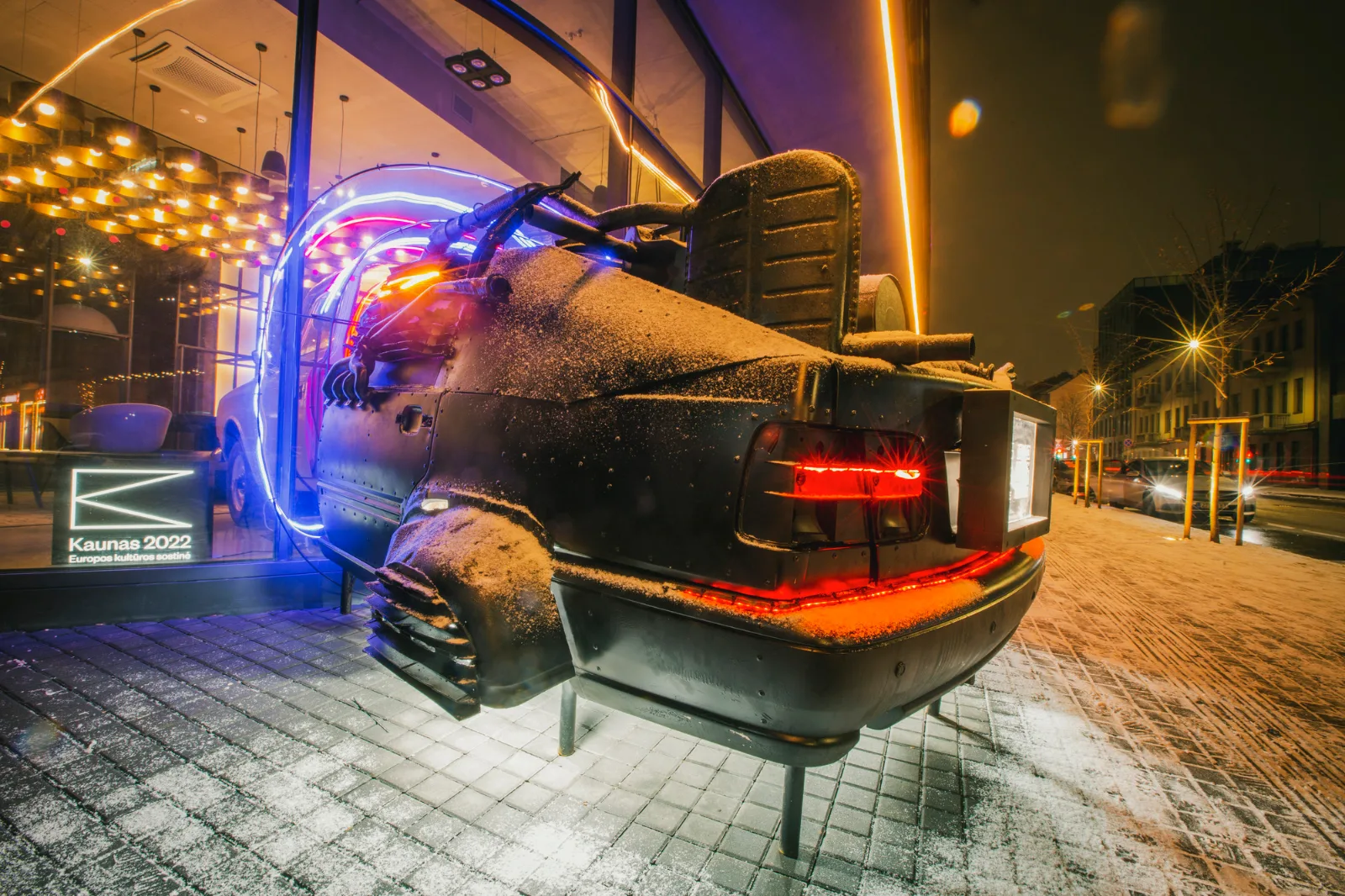 Kosmos Car – the rear of a car with neon lights outside a glass-fronted building at night