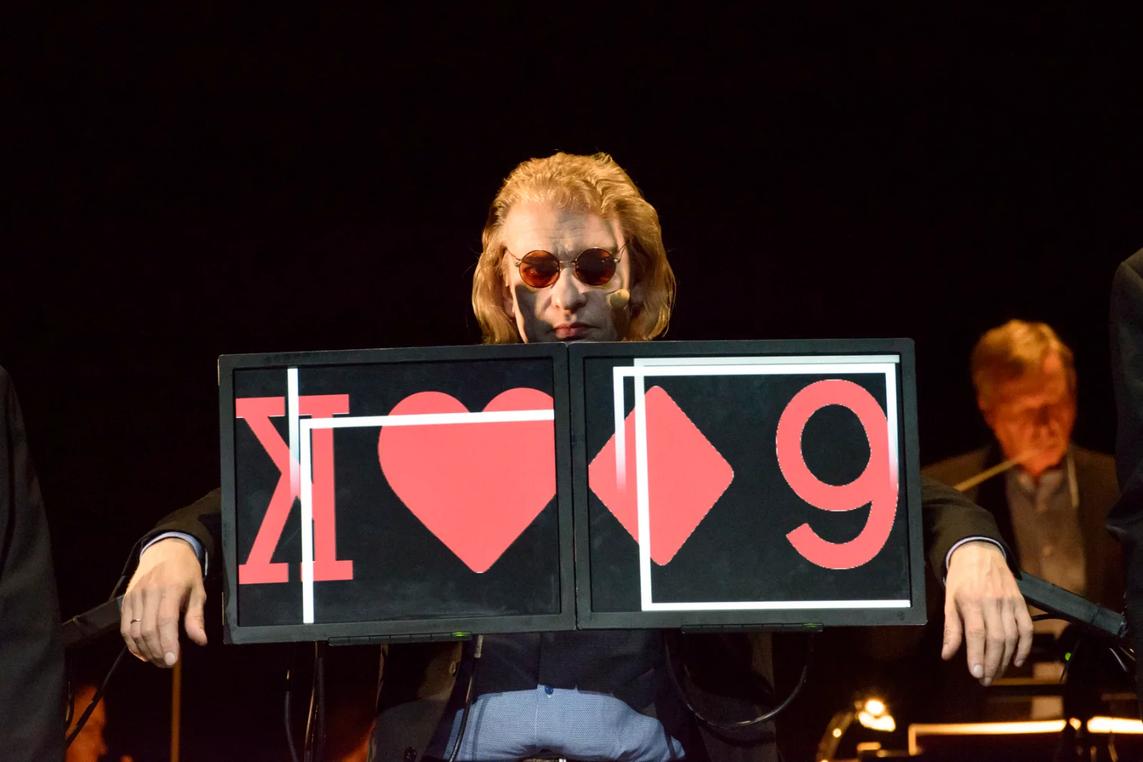 The Man Who Mistook His Wife For a Hat – a singer in sunglasses holds up a card with playing-card symbols