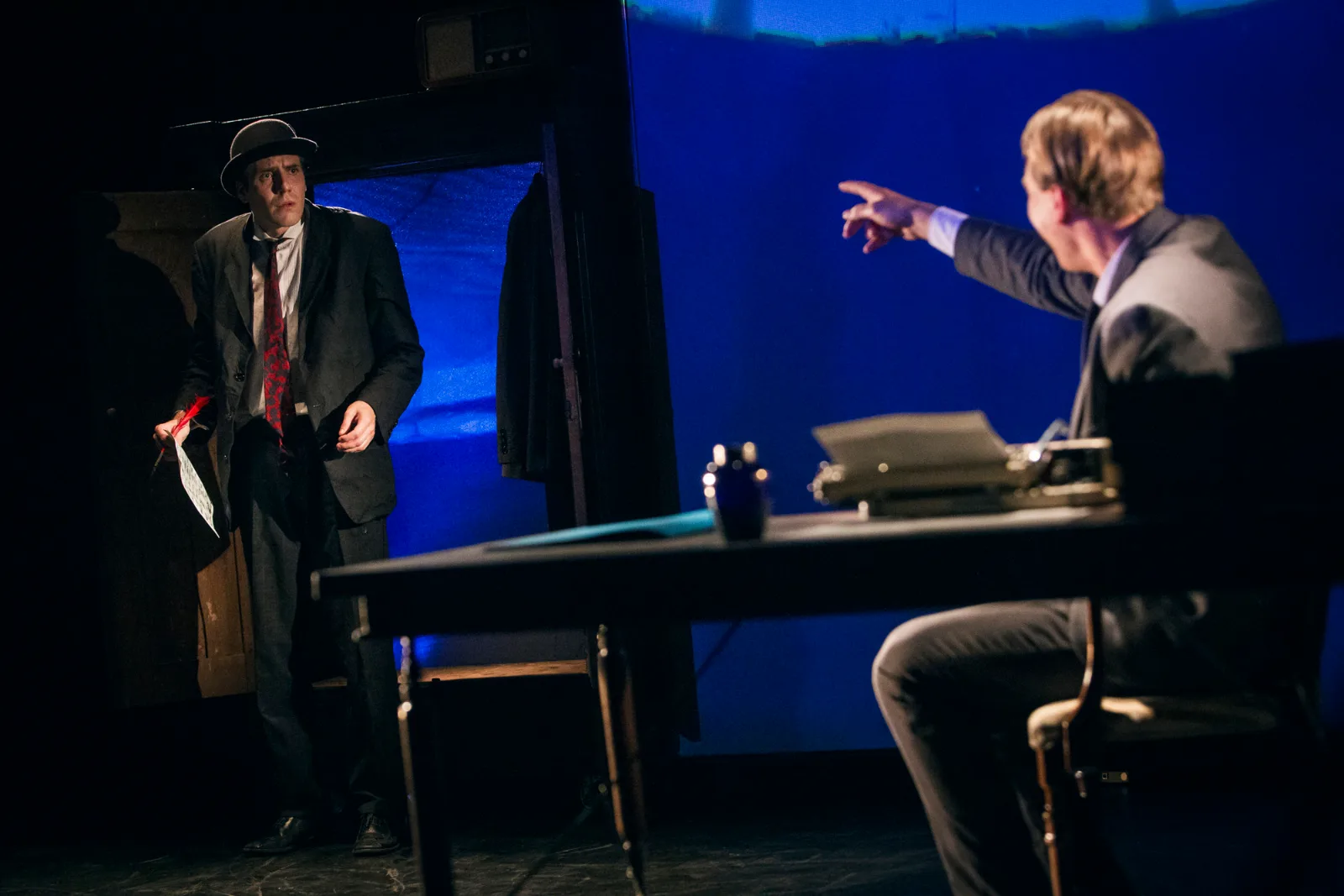 Kafka Insomnia – an actor in a coat and hat faces a man pointing from behind a desk