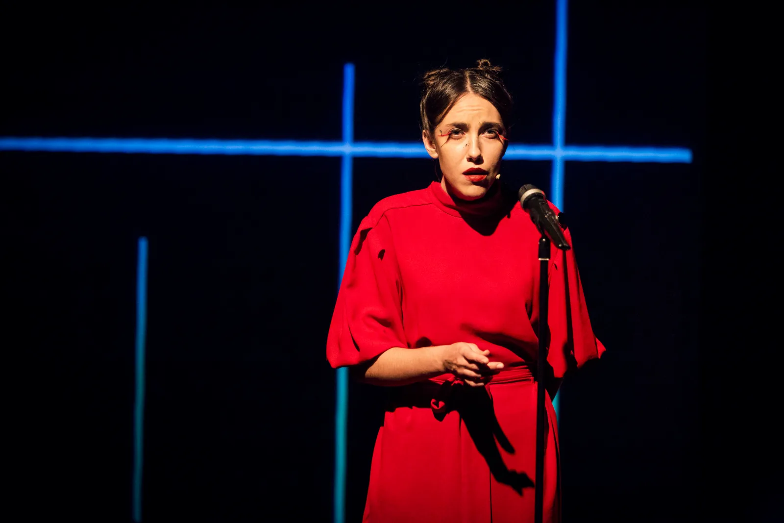 Kafka Insomnia – a woman in a red dress speaks into a microphone among lines of blue light