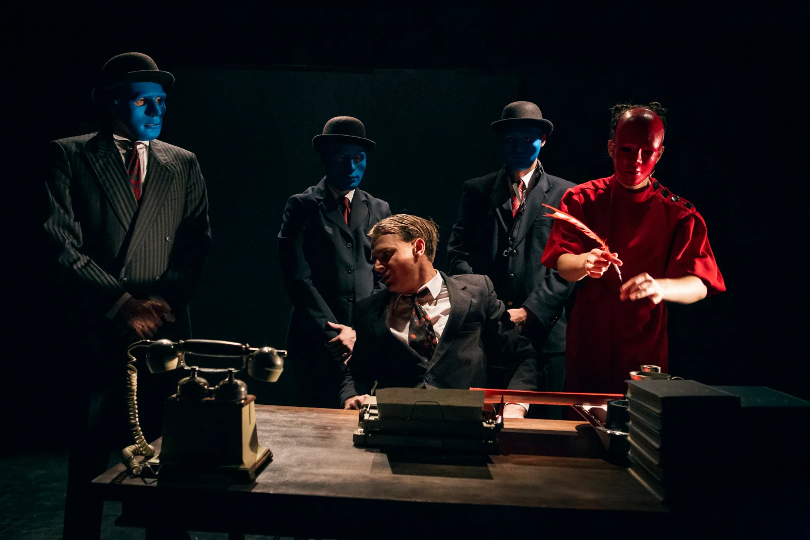 Kafka Insomnia – figures with blue and red faces in bowler hats surround a man at a desk