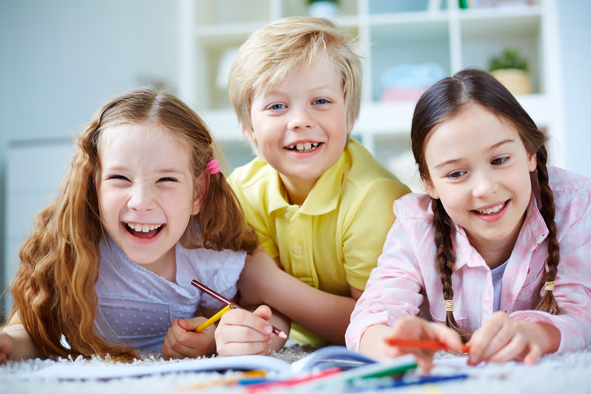 Three children lying on the floor smiling and drawing in a book with colored pencils, two girls with braids and a boy in a yellow shirt between them.