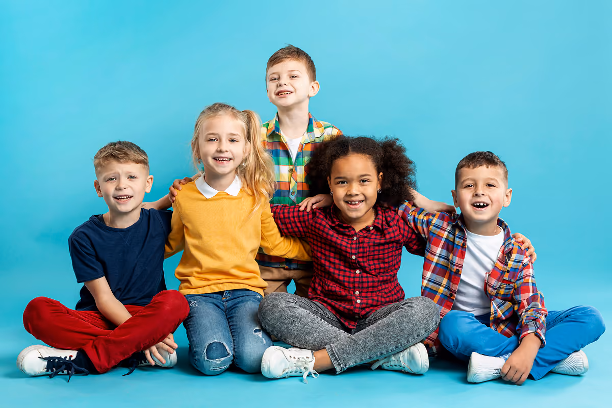 Five smiling children sitting on the floor against a blue background, with arms around each other, wearing casual colorful clothes.