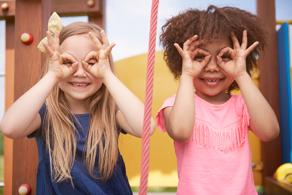 Two young girls smiling and pretending to wear glasses with their hands in a playful gesture at a playground.