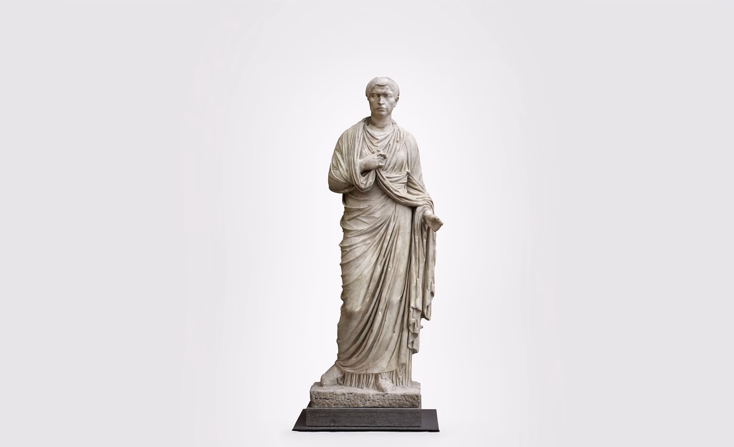 Marmorstatue af en romersk kvinde iført en foldet toga midtstillet på en base med et neutralt gråt baggrund.