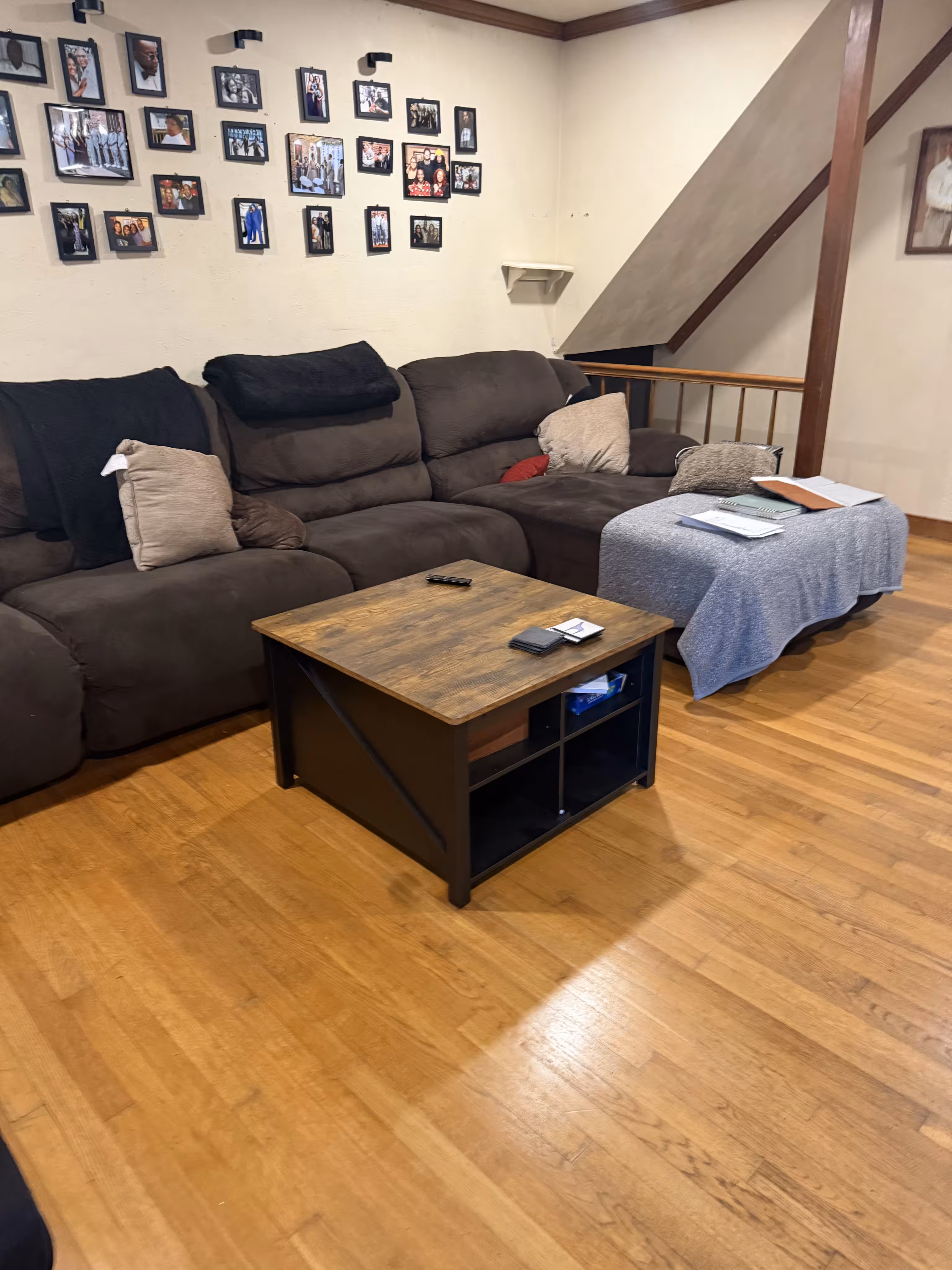 Living room with a brown sectional sofa adorned with beige and black pillows, a wooden coffee table with remote controls and cards, and a blue-gray throw covering a side table holding papers, all set on a hardwood floor with a wall decorated with numerous framed photos.