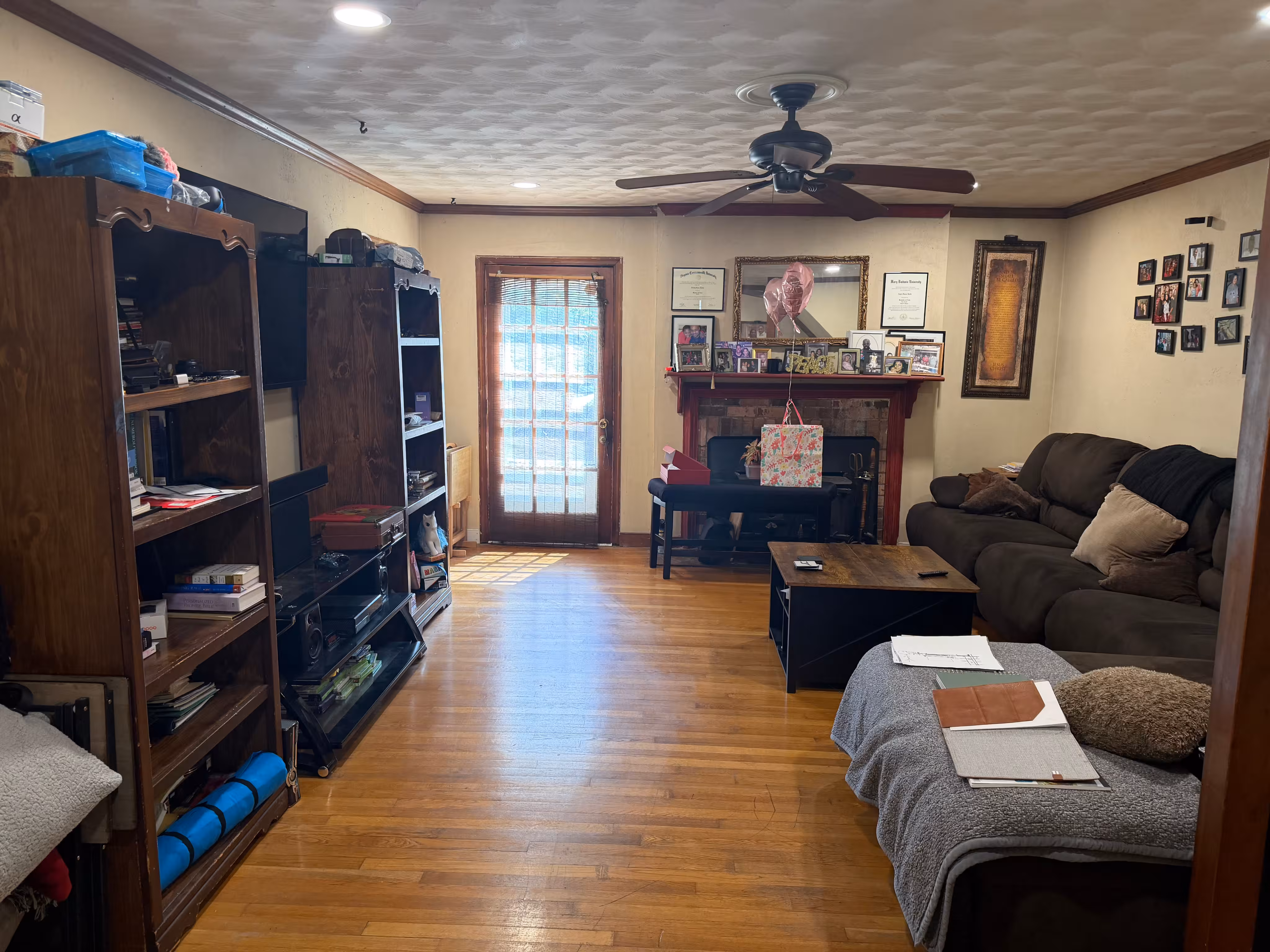 Living room with wooden floor, brown ceiling fan, bookshelves on the left, a dark brown sofa and a coffee table on the right, and a fireplace with picture frames, balloons, and a gift bag in the background near a glass door with a curtain.