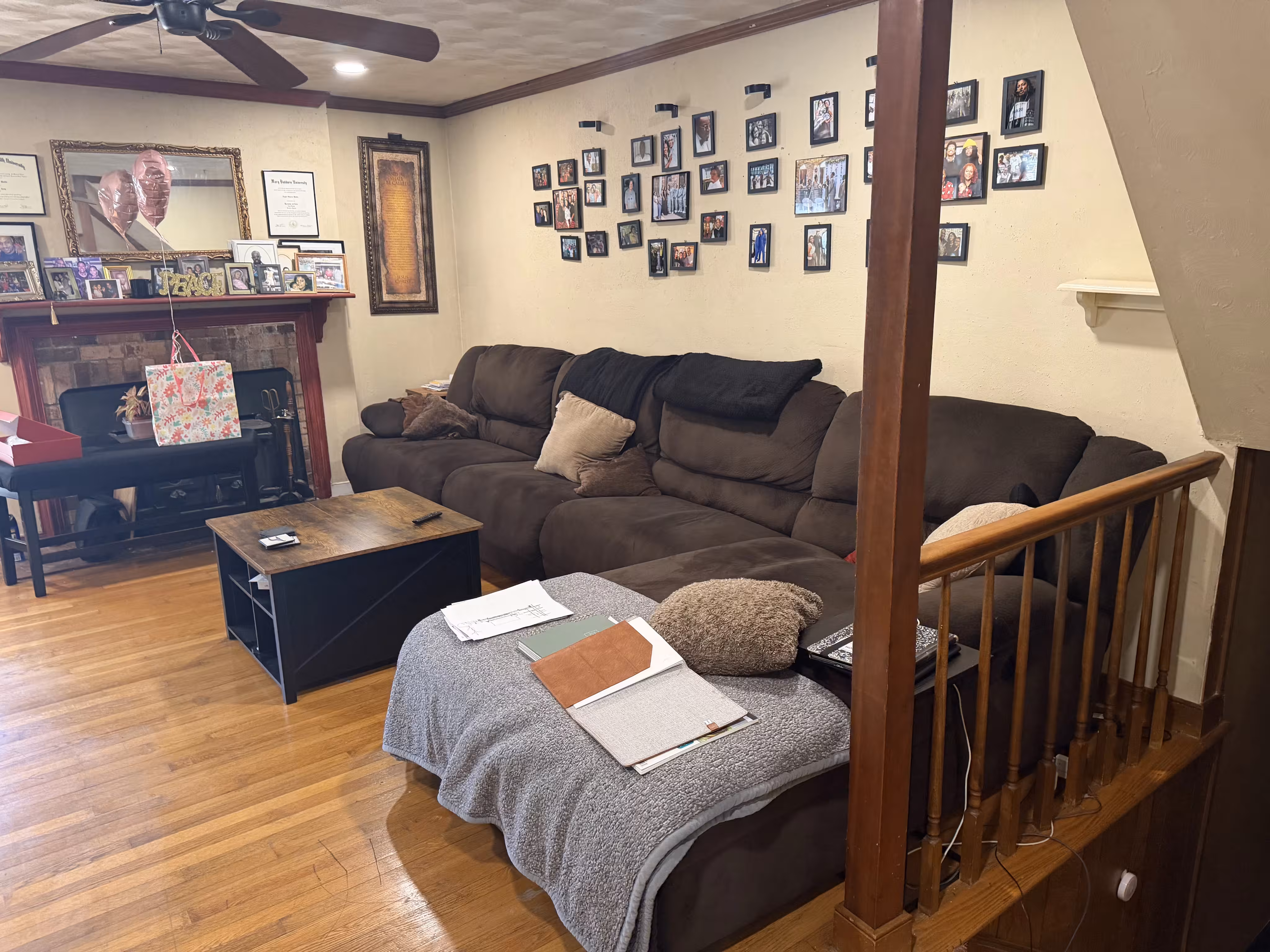 Cozy living room with a large brown sectional sofa covered partly with a gray throw and papers, a wooden coffee table, hardwood floor, and a wall decorated with numerous framed photos above the sofa; a fireplace with balloons and framed certificates is in the background.