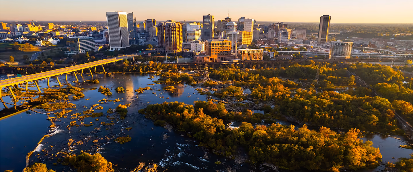 Aerial view of a city skyline at sunset with a river running through a large park area filled with green and orange autumn trees and a bridge crossing the river.