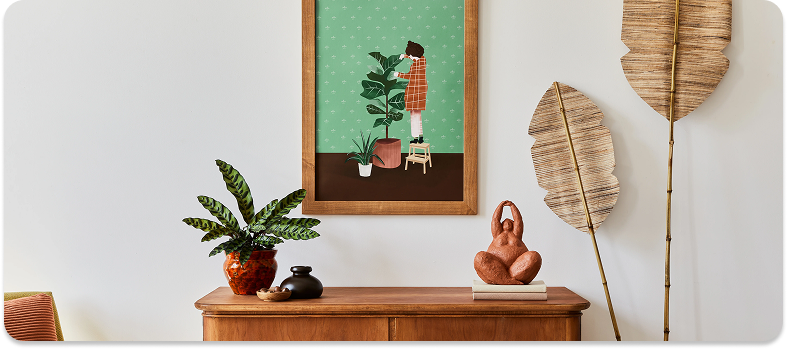 A wooden sideboard with a green leafy potted plant, two small decorative jars, and a terracotta sculpture of a seated figure on top; above it hangs a framed illustration of a person in an orange coat tending a houseplant; two large decorative dried palm leaves lean against the white wall on the right.