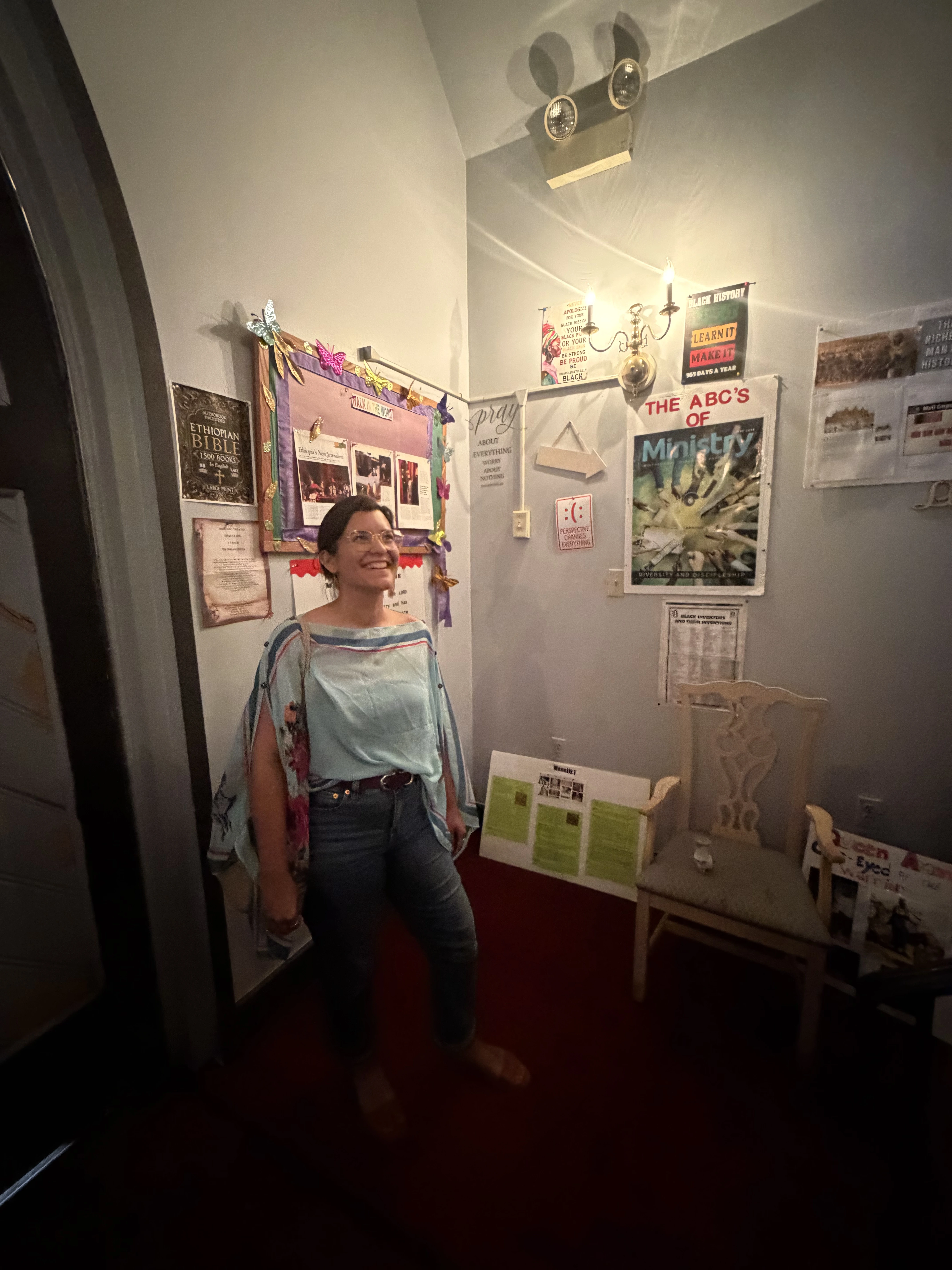 A woman wearing glasses, a light blue top with floral patterns, and blue jeans stands smiling inside a room with walls covered in colorful posters and bulletin boards, including one titled 'THE ABC’S OF Ministry'. There is a wooden chair and various papers displayed around her. The room is softly lit by a wall-mounted light fixture with two candles.