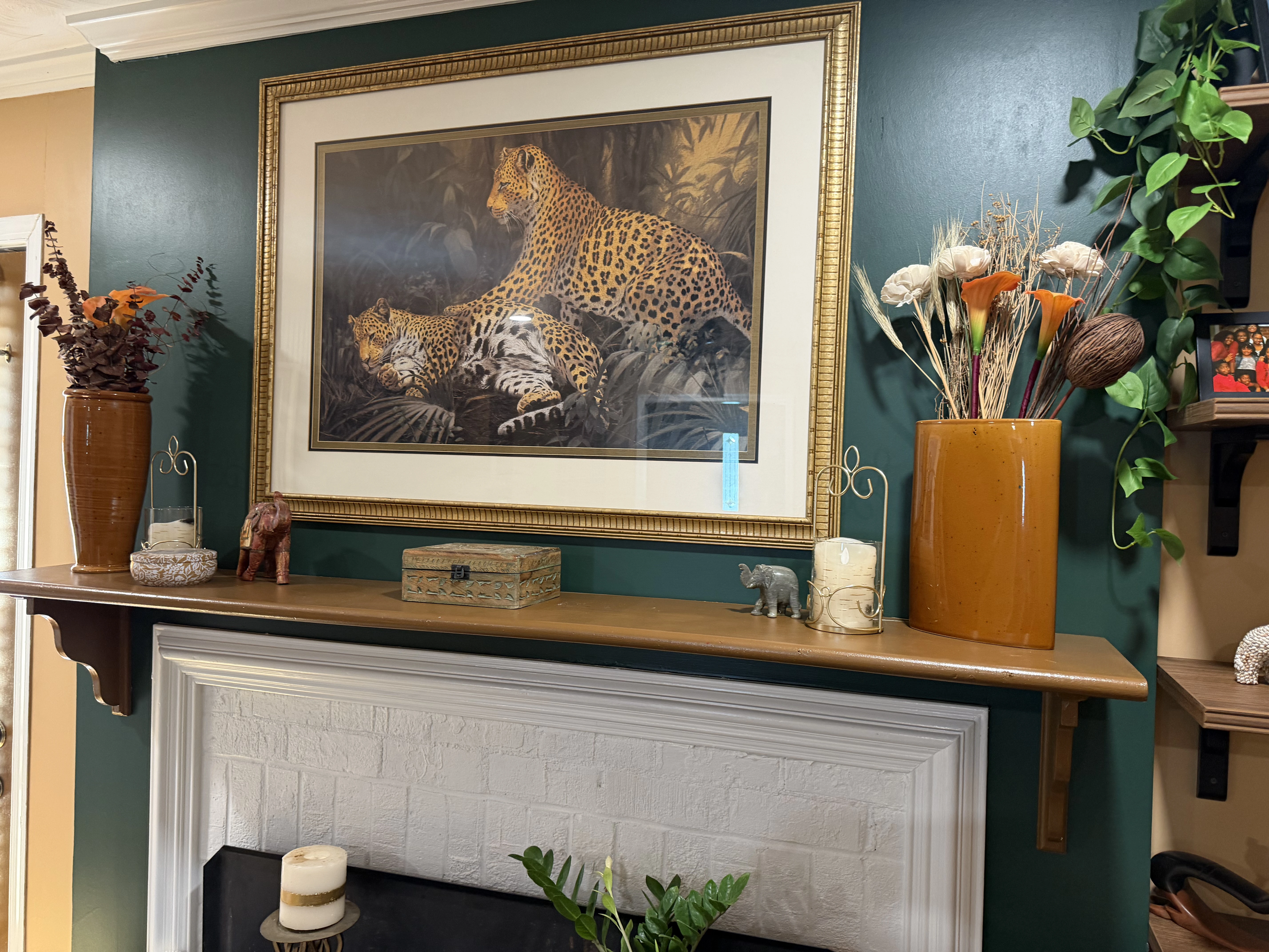 Wall-mounted wooden shelf above a white painted fireplace mantel displaying a framed painting of two leopards in a forest, two large orange vases with floral and dried plant arrangements, candles in holders, a small carved wooden elephant, a decorative box, and a small grey elephant figurine, with green wall paint and part of a wooden shelf with plants on the right.