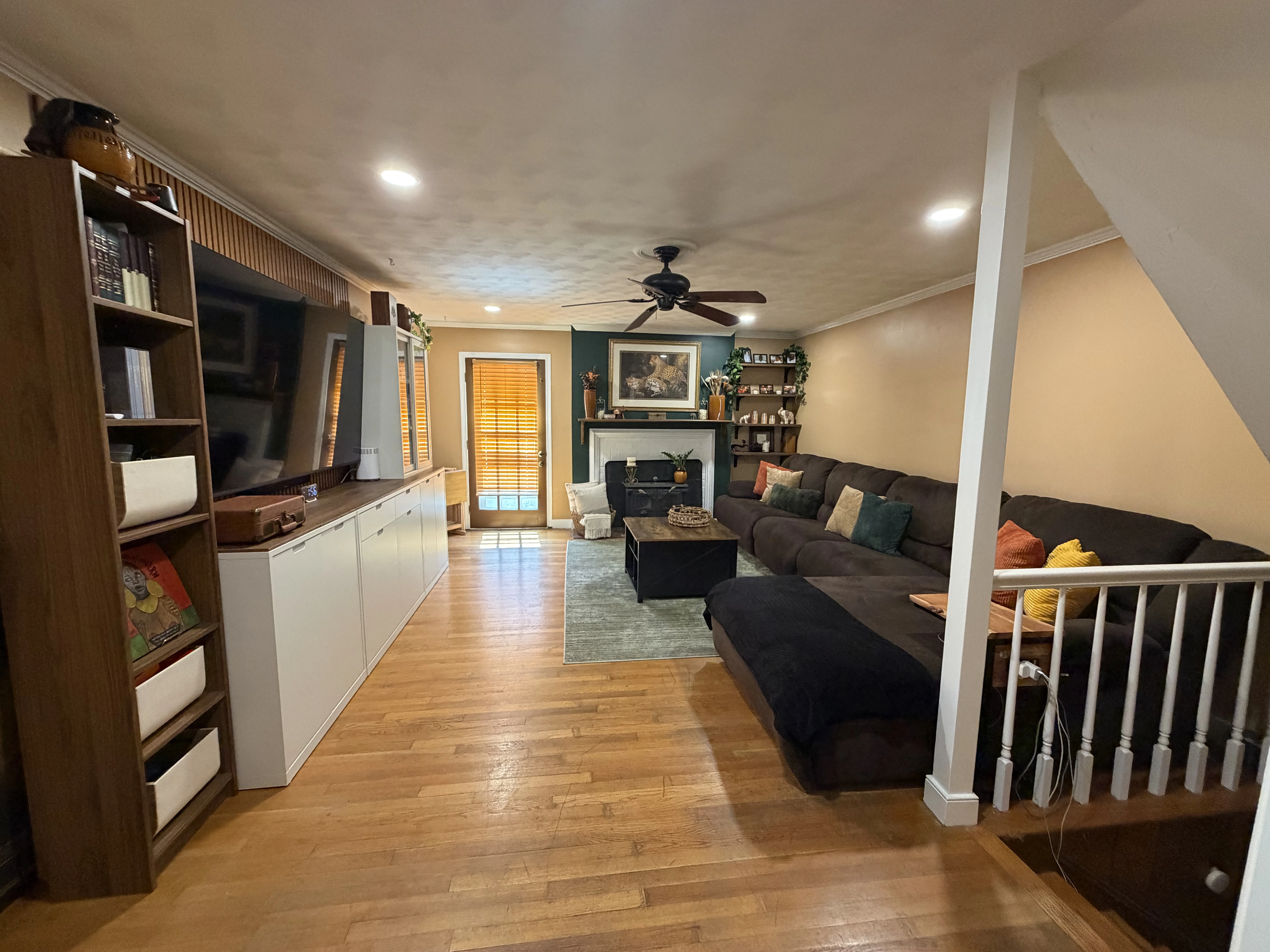 Living room with a large dark sectional sofa adorned with colorful pillows, wooden floor, white cabinets and shelves on the left, a fireplace with a leopard painting above, ceiling fan, and a door with wooden blinds letting in light.