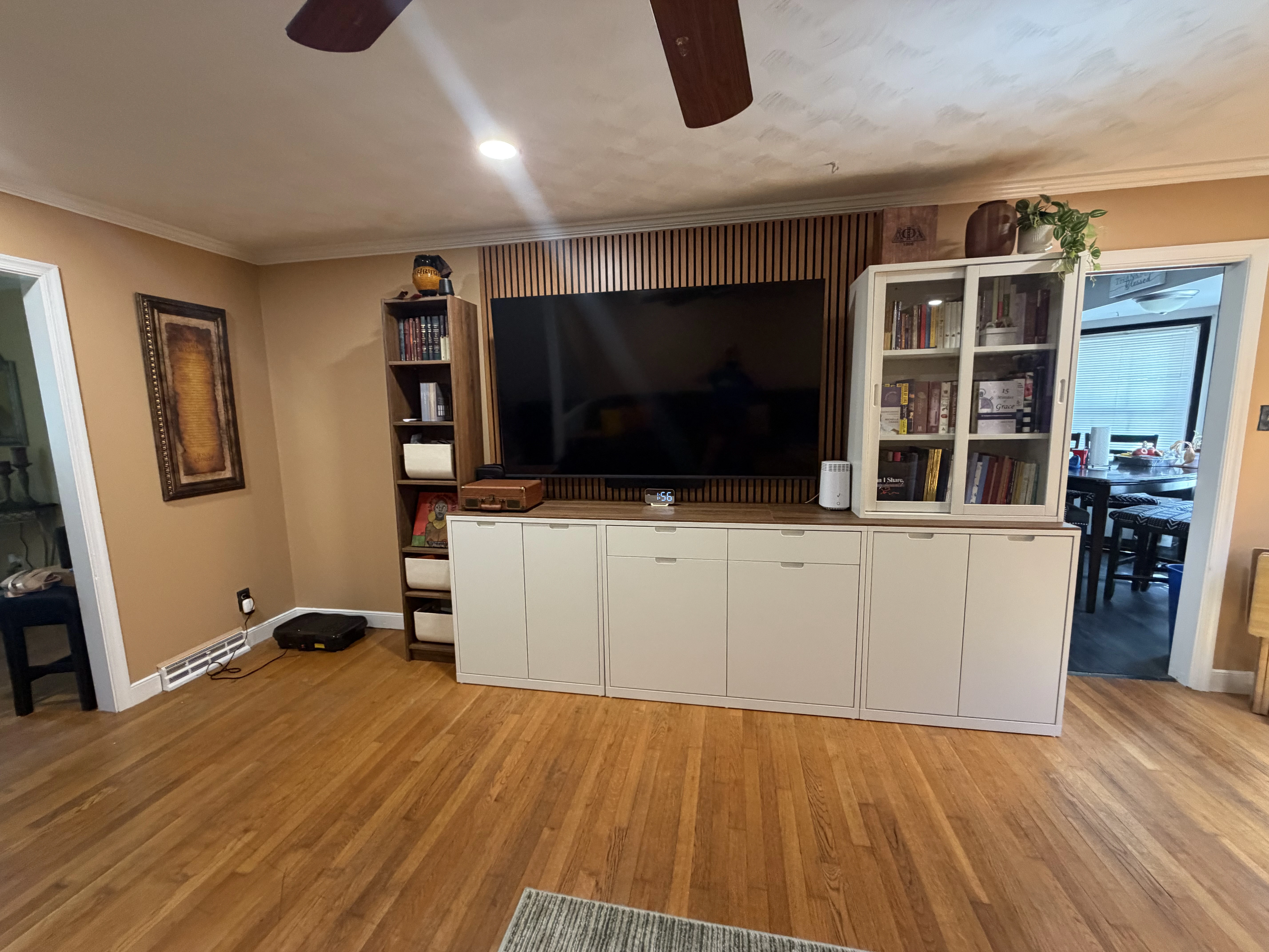 Living room with wooden floor and beige walls featuring a large flat-screen TV mounted on a wooden slatted panel. Below the TV is a white cabinet with multiple compartments. To the left of the TV is a tall wooden shelf with books and decorative items. To the right is a white glass-door cabinet with books and plants on top. Two doorways on either side reveal adjacent rooms, one with a dark chair and table and the other with a dining table and chairs.