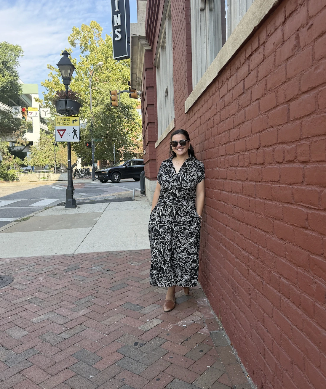 Smiling woman wearing sunglasses and a black dress with a white floral pattern leaning against a red brick wall on a sidewalk in an urban area with trees and street signs in the background.