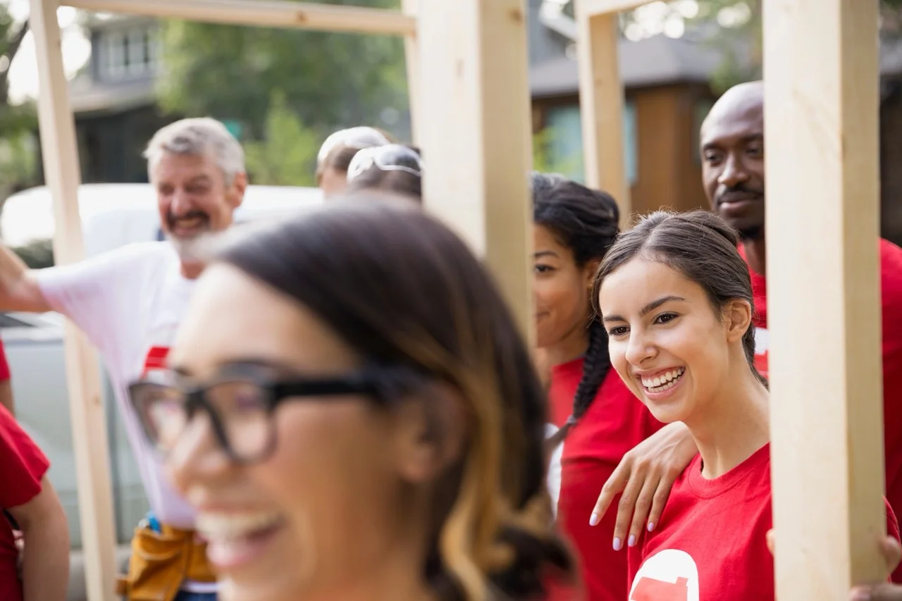 Group of diverse volunteers in red shirts smiling and assembling wooden frames outdoors in a community building project.