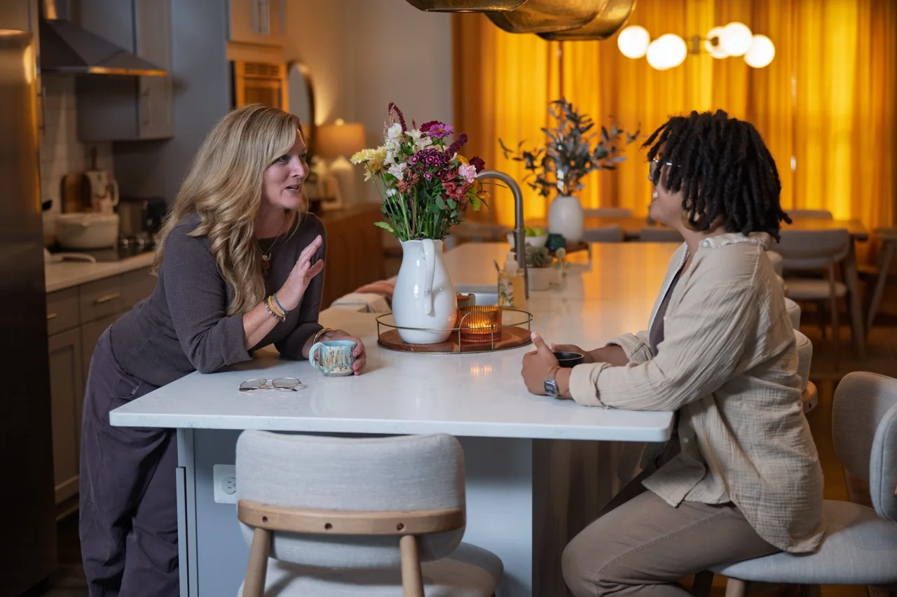 Two women are sitting at a kitchen island table having a conversation; one woman with long blonde hair is leaning forward holding a mug, the other with curly dark hair and glasses is seated holding a small cup; there is a white vase with colorful flowers and a lit candle on the table, with warm yellow curtains and string lights in the background.