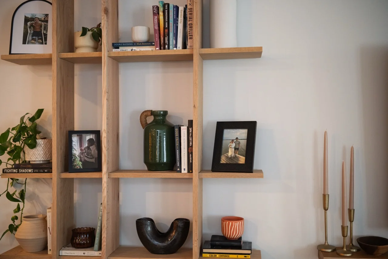 Wooden shelving unit against a light wall featuring decorative items including potted plants, framed photos, books, a green vase, a black decorative piece, a small orange cup, and four tall pink candles in brass holders.