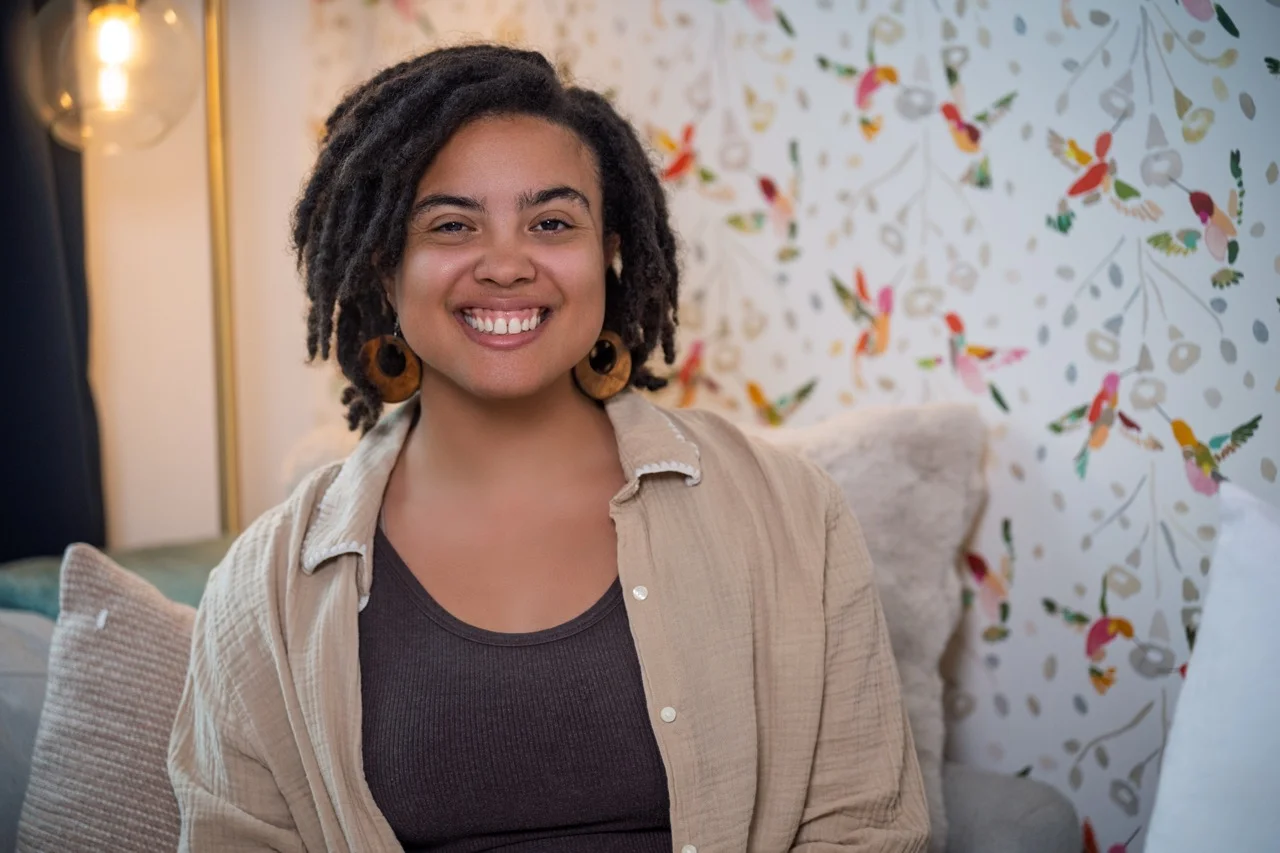 Smiling woman with natural curly hair, wearing a beige button-up shirt over a dark tank top, sitting on a couch with patterned cushions and a floral wall behind her.