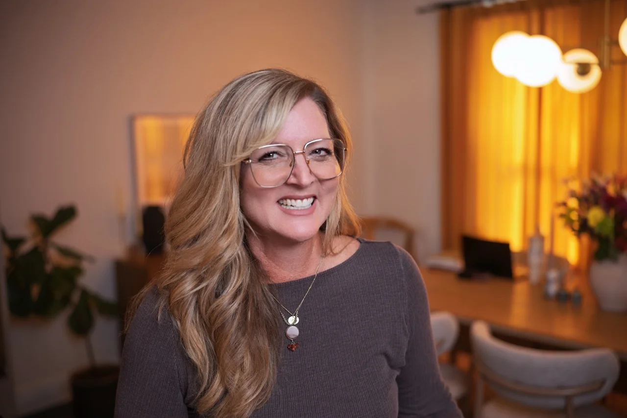 Smiling woman with long blonde hair wearing large glasses and a purple top standing in a warmly lit room with a wooden table, chairs, a plant, and hanging lights.