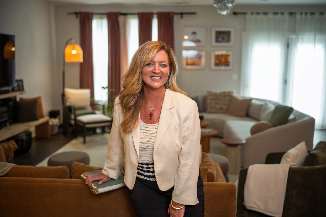A smiling woman with long blonde hair wearing a white blazer and striped top leans on the back of a brown couch in a warmly lit living room with modern furniture, including a TV, armchair, sofa, and framed pictures on the wall.