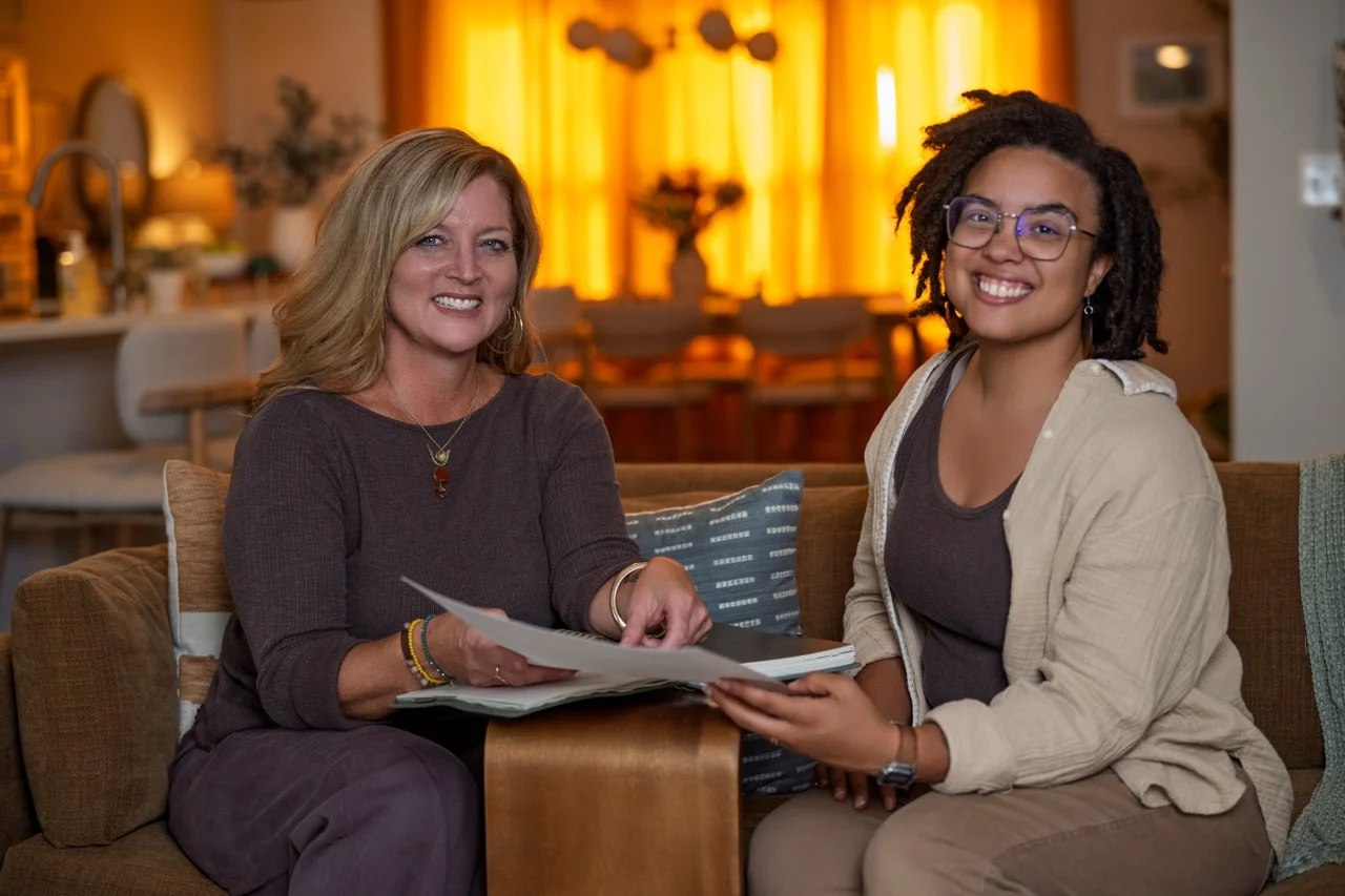 Two women sitting on a brown couch in a warmly lit room, smiling and holding open a book or album together while looking at the camera.