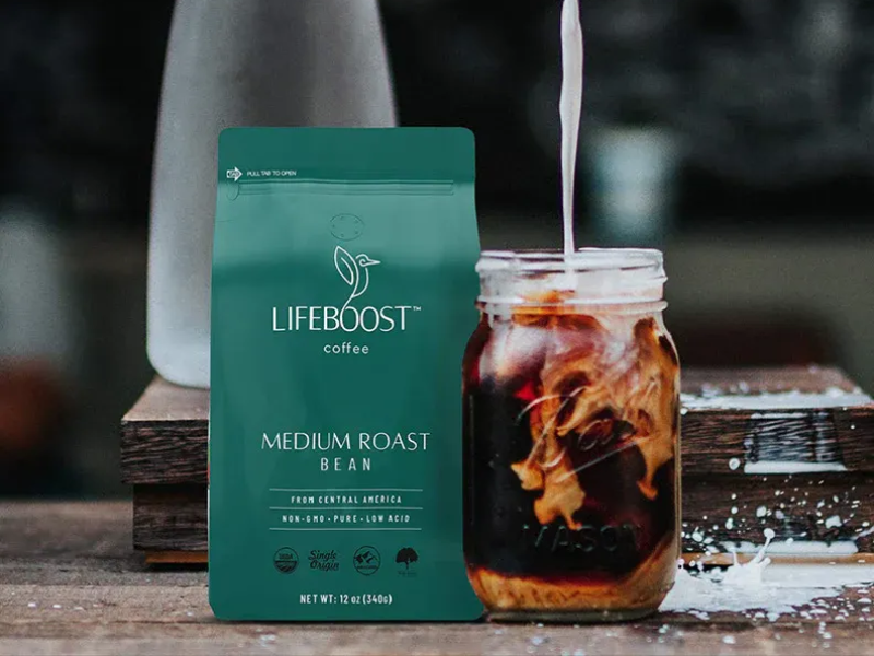 A teal bag of LifeBoost coffee labeled Medium Roast Bean next to a mason jar filled with iced coffee and cream being poured into it on a wooden surface.