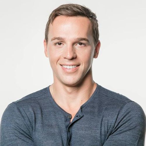 Smiling young man with short brown hair wearing a dark gray henley shirt, posing with arms crossed against a plain white background.