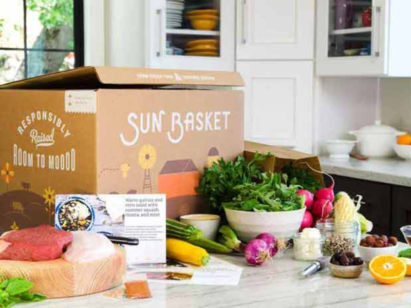 Kitchen countertop with unpacked Sun Basket meal kit showing a cardboard box, fresh raw meats on a wooden cutting board, a bowl of leafy greens, various fresh vegetables including radishes and squash, jars with seeds and other ingredients, and a halved orange.